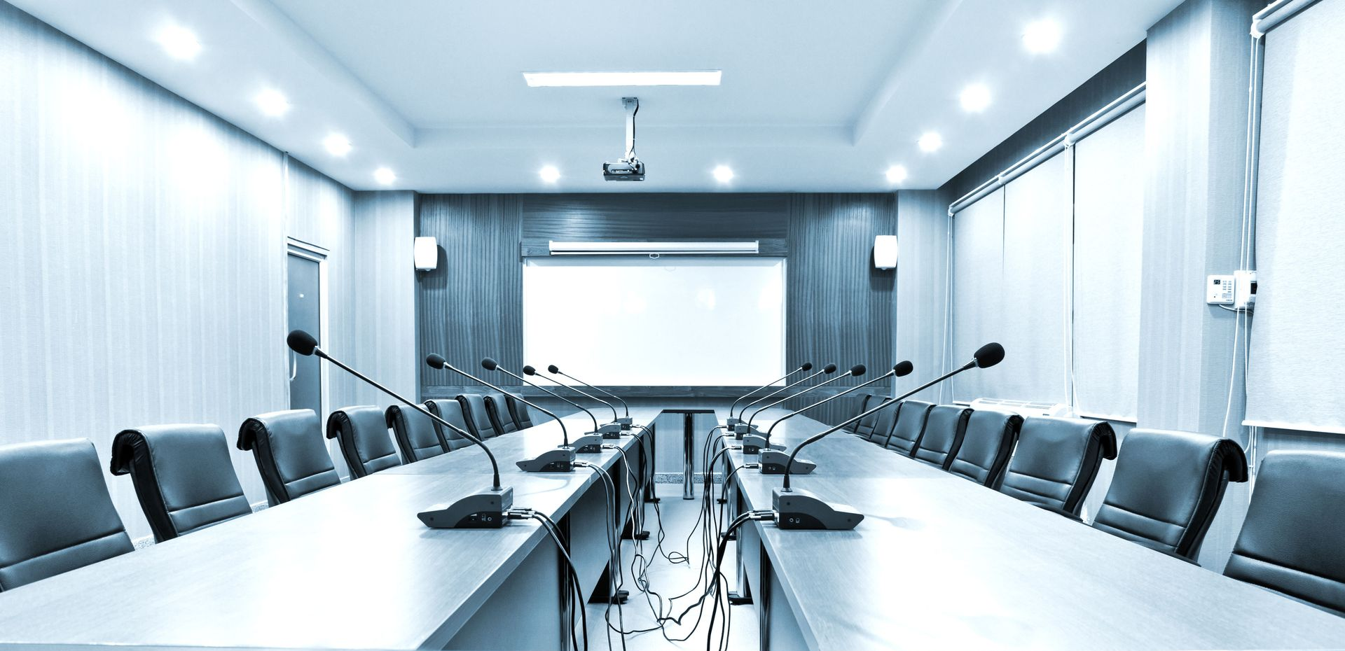 Empty conference room with long table, microphones, chairs, and projector screen.