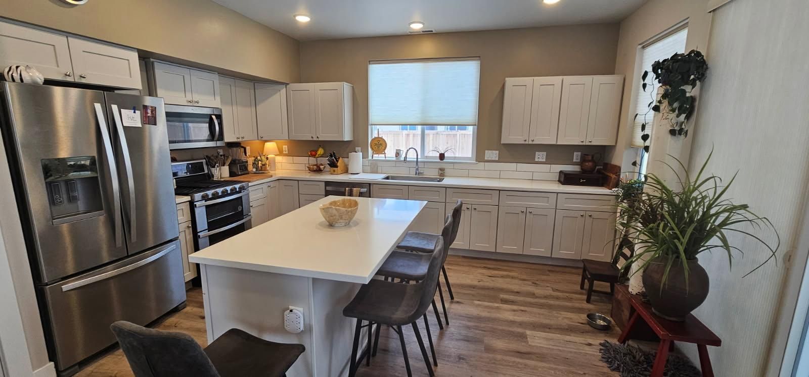 A modern kitchen with white cabinets, stainless steel appliances, a large island, and a window. Wood flooring.