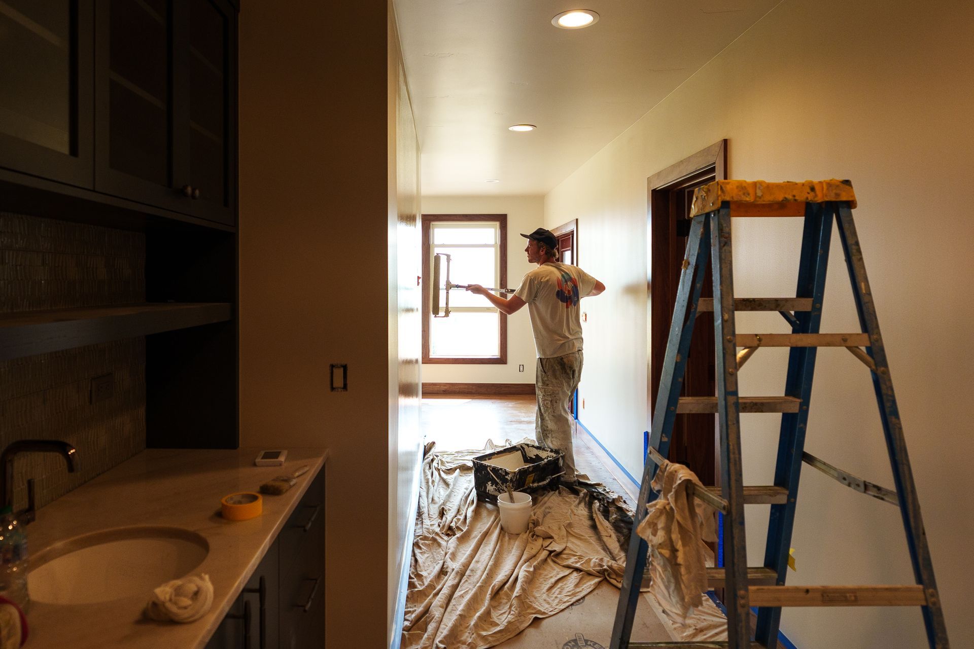 A person paints a hallway, standing on a ladder. Paint bucket and drop cloth on the floor.