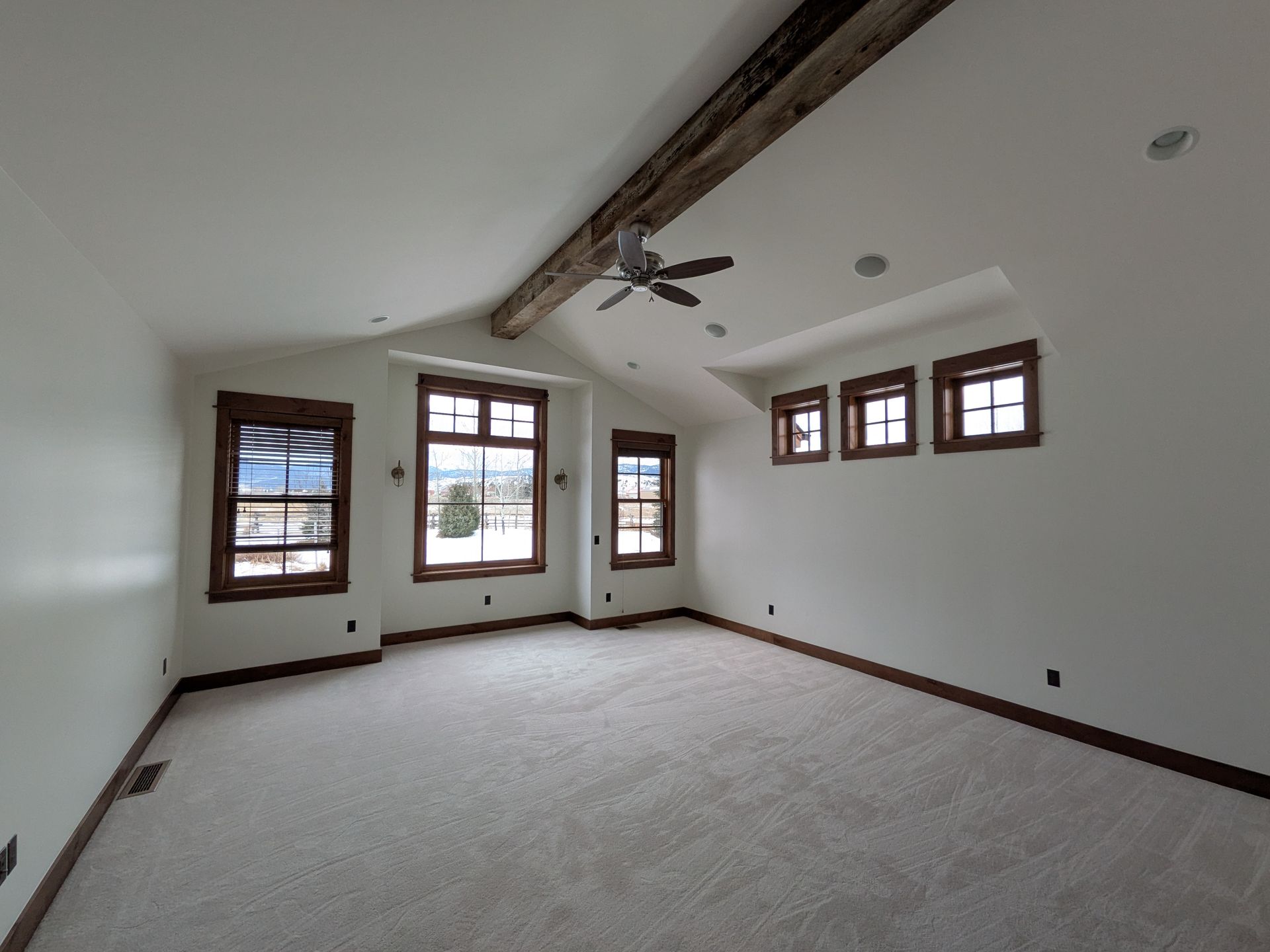 Spacious, empty bedroom with multiple windows, wooden beams, light carpet, and white walls.