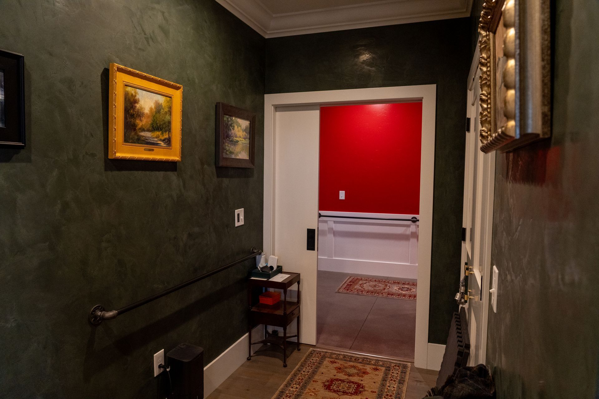 Dark green hallway with framed art, open white door leading to a room with red wall and rug.