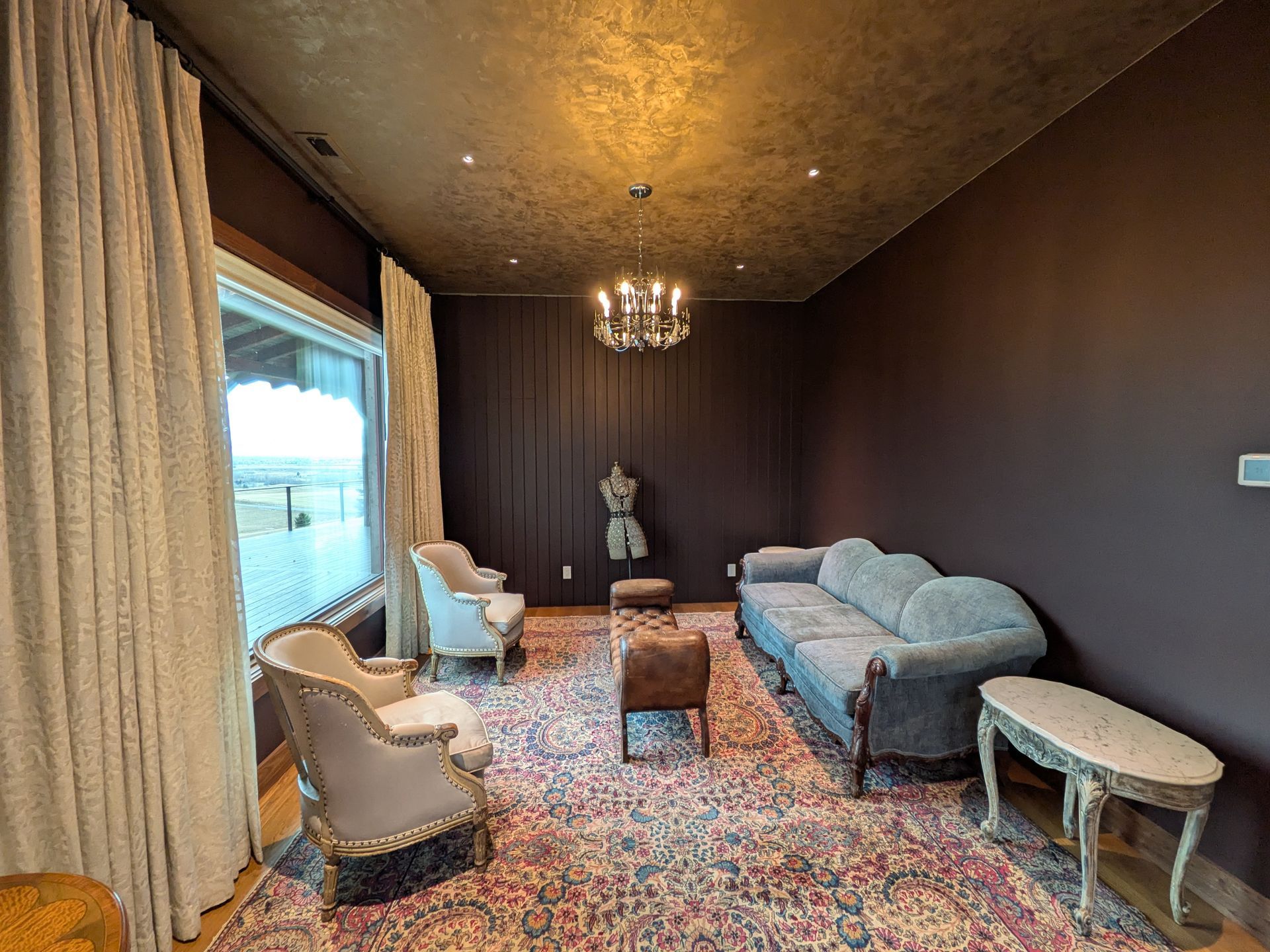 Elegant room with velvet sofa, armchairs, ornate rug, large window, and chandelier. Dark walls and golden ceiling.