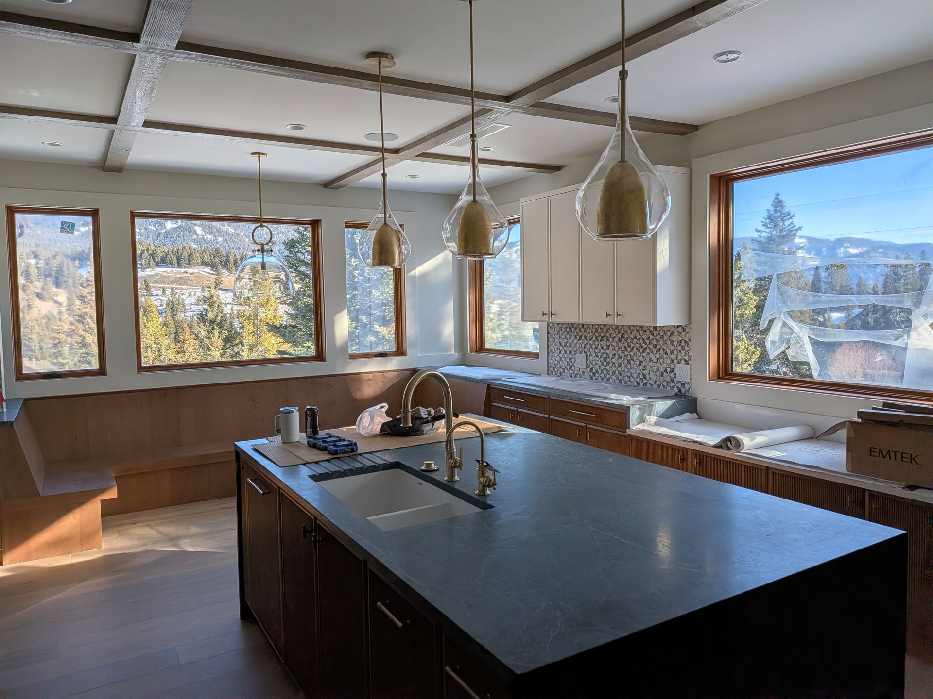 Kitchen with island, light fixtures, and windows overlooking a snowy landscape. Brown cabinets, white walls, and a wood beam ceiling.