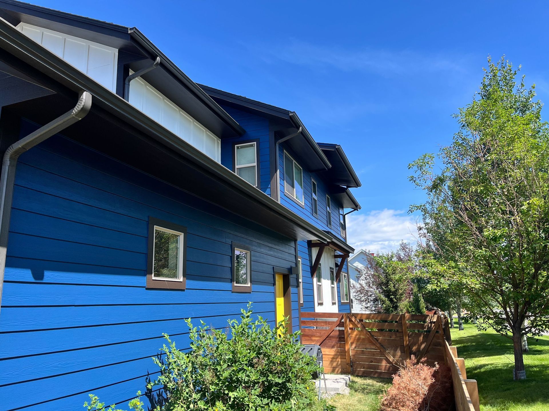 Blue house with black trim and yellow door, under a bright blue sky with a green tree and brown fence.