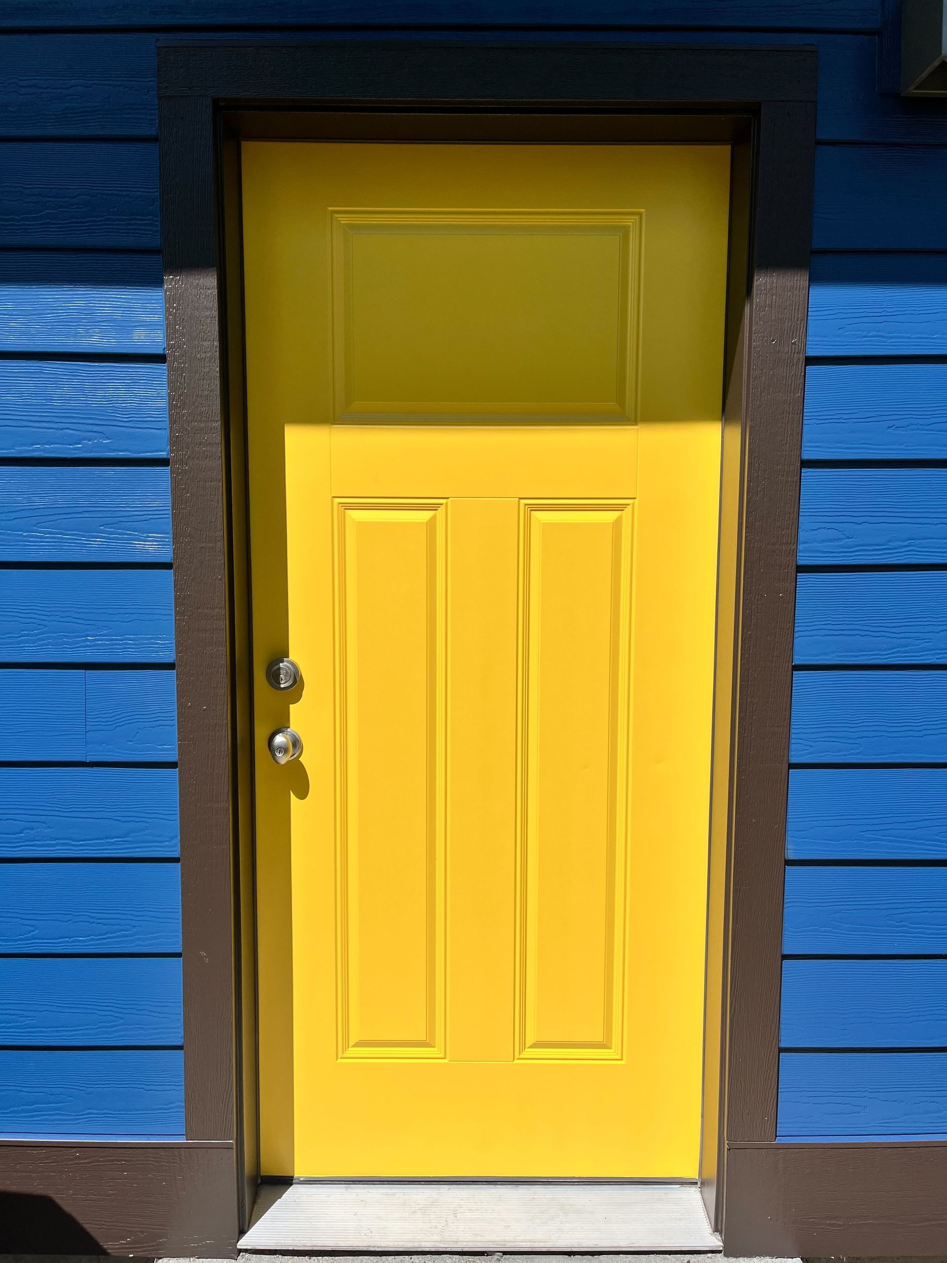 Yellow door with brown trim in a blue building. Sunlight casts shadows.