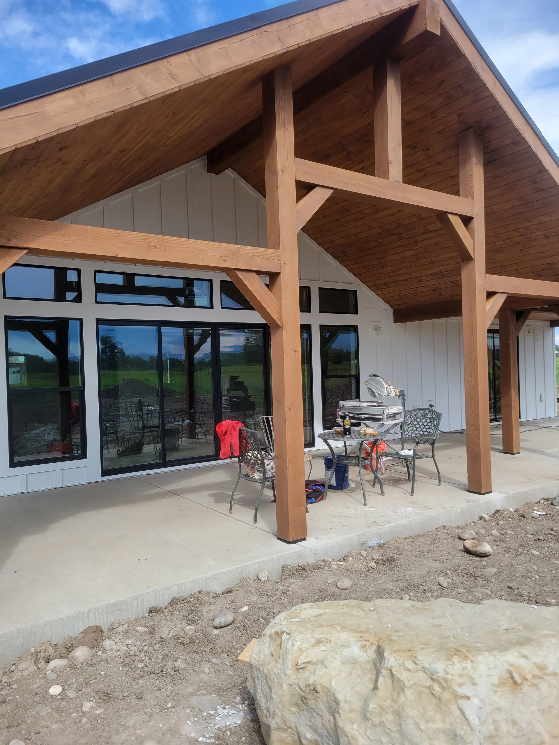 A modern white house with dark trim and a wooden porch is shown. Outdoor furniture sits on a concrete patio.