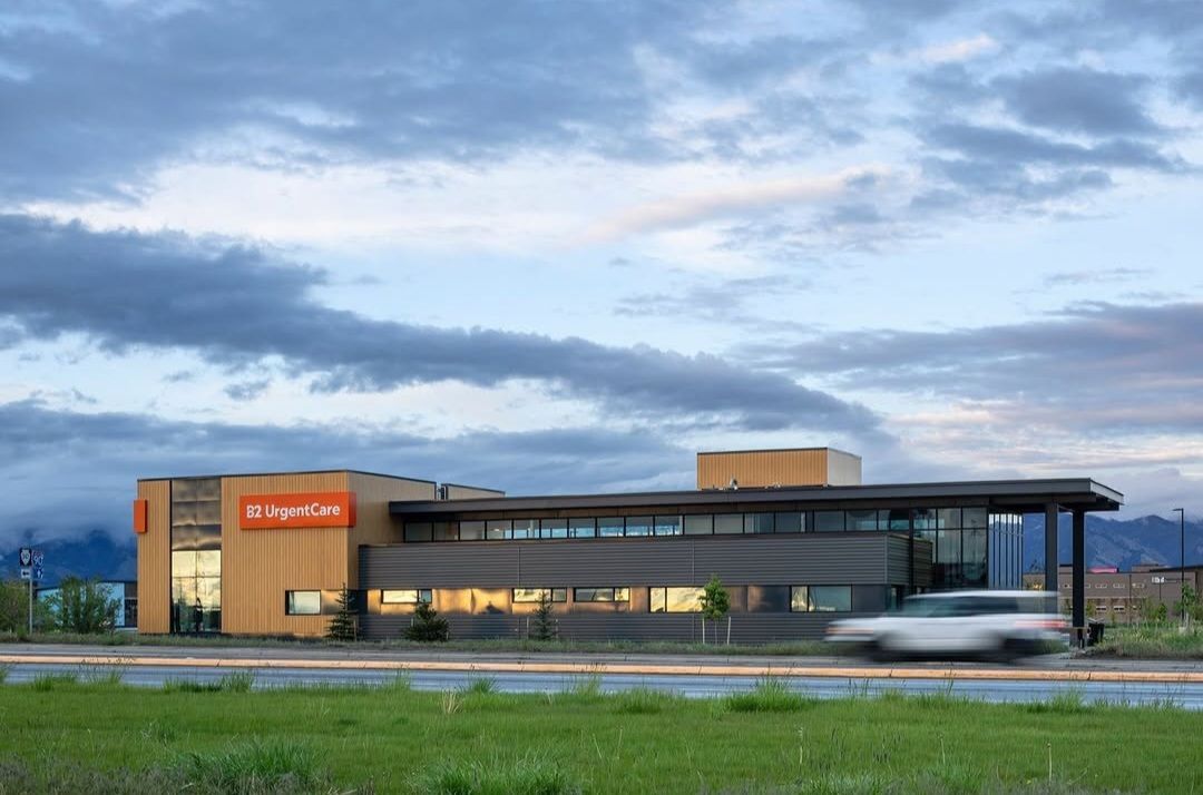 Modern, two-story building with orange signage, wooden panels, and glass windows. A blurred car drives past the building on a road with a green field and cloudy sky.