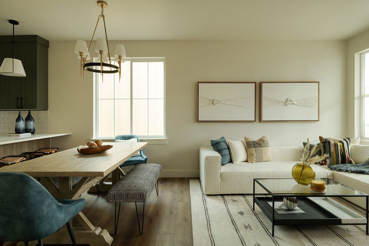 A modern living and dining room with a neutral color palette. Features a white couch, wooden table, and a dark green kitchen.