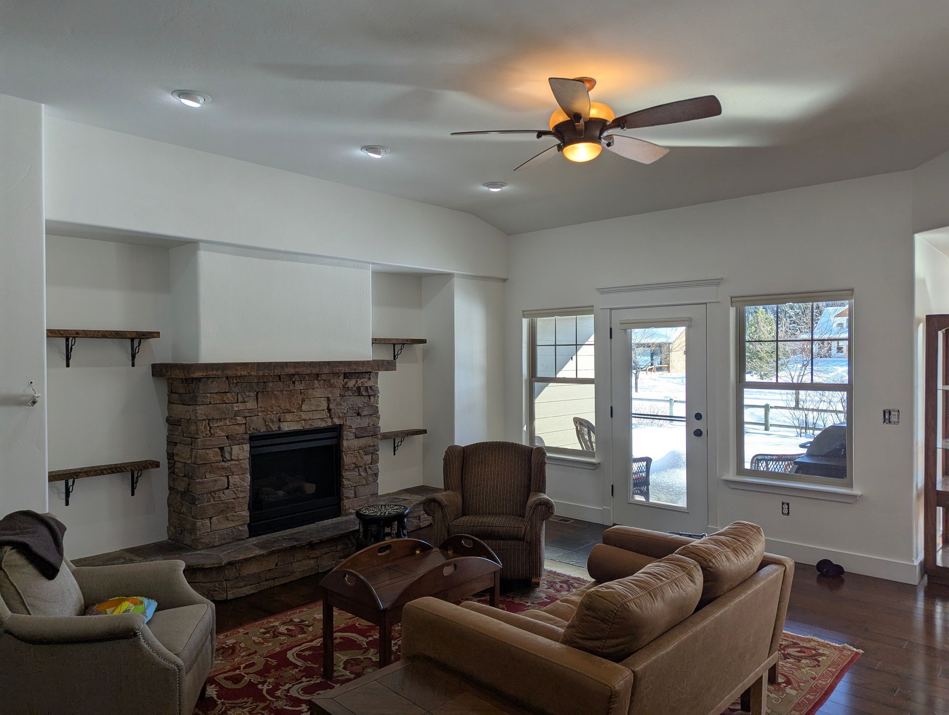 Living room with stone fireplace, built-in shelves, and a ceiling fan. Two armchairs and a sofa face the fireplace. A doorway leads to outside.