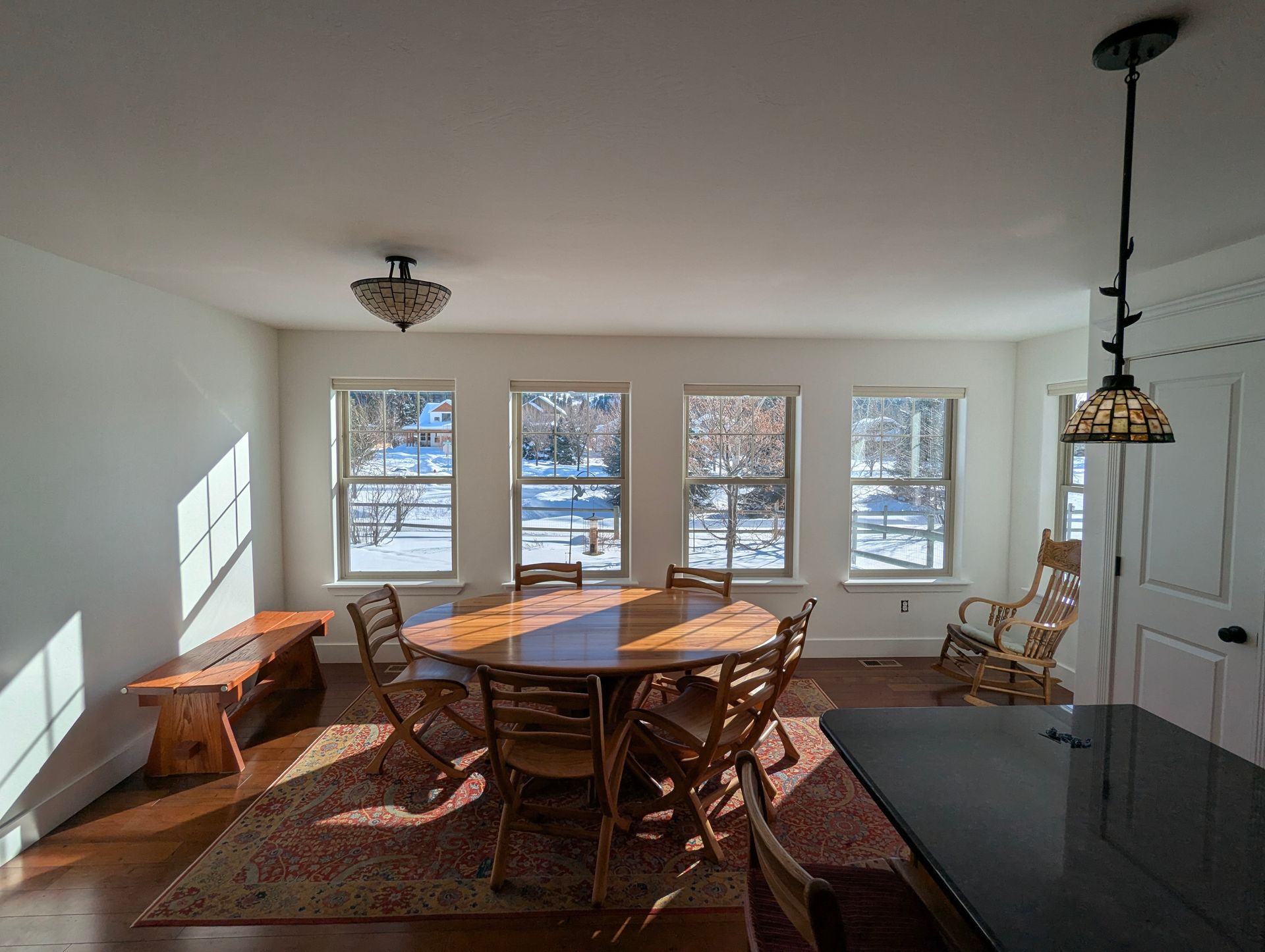 Dining room with a wooden table and chairs, a bench, and a rug on a hardwood floor. Sunlight streams through three windows with a snowy view.
