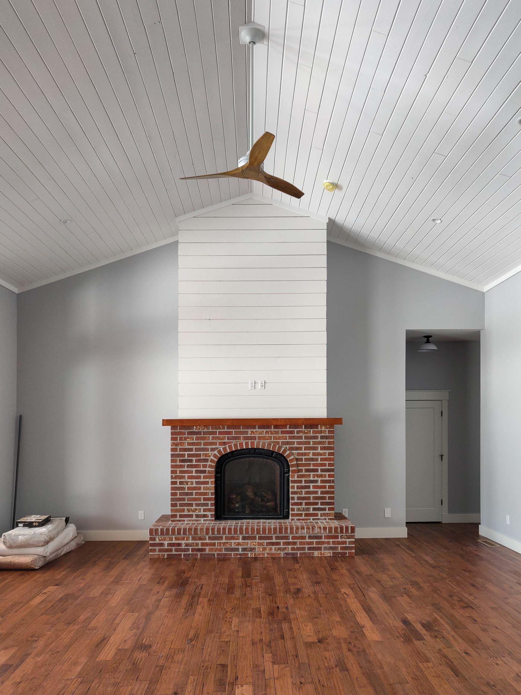 Living room with brick fireplace, wooden floor, and white ceiling with fan. Gray walls and doorway.