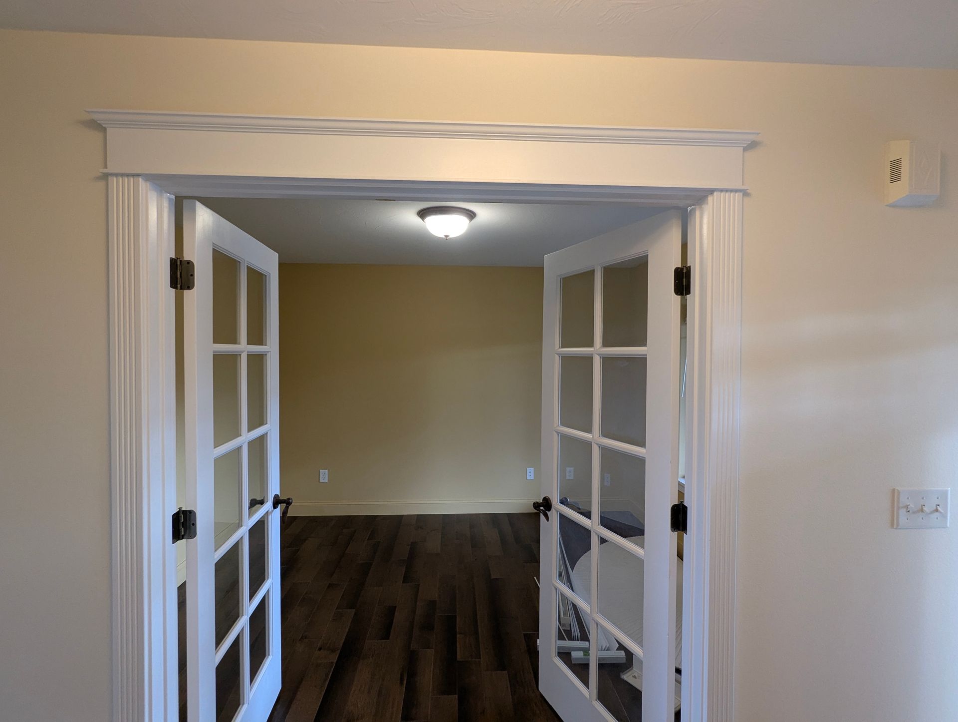 White French doors open into a room with tan walls and dark wood flooring. Cream-colored trim surrounds the doorframe.