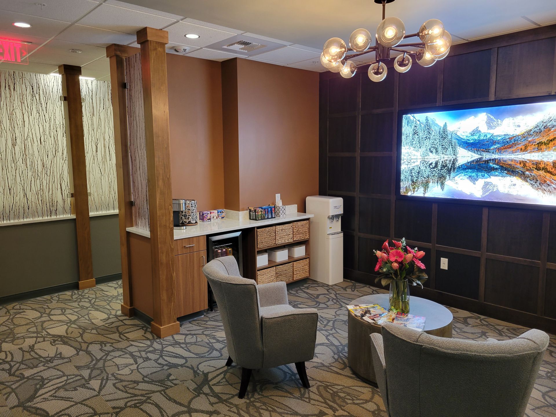 Cozy waiting area with armchairs, coffee station, and large TV screen displaying a scenic landscape. Warm lighting and neutral color palette.