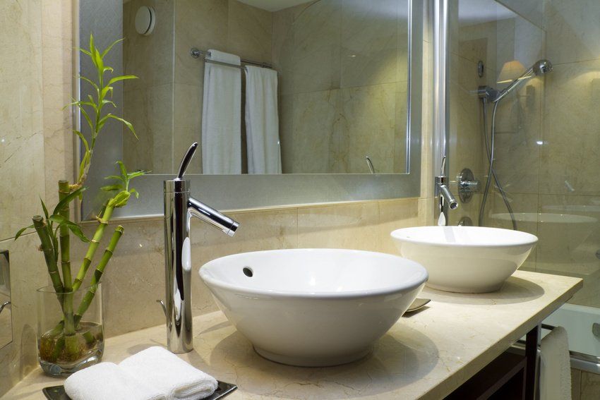 Modern bathroom with two white bowl sinks on a beige countertop, a large mirror, and bamboo in a vase.