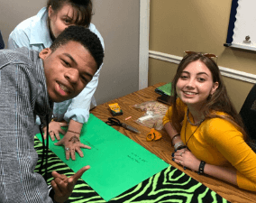 A group of people are sitting at a table with green paper and scissors.