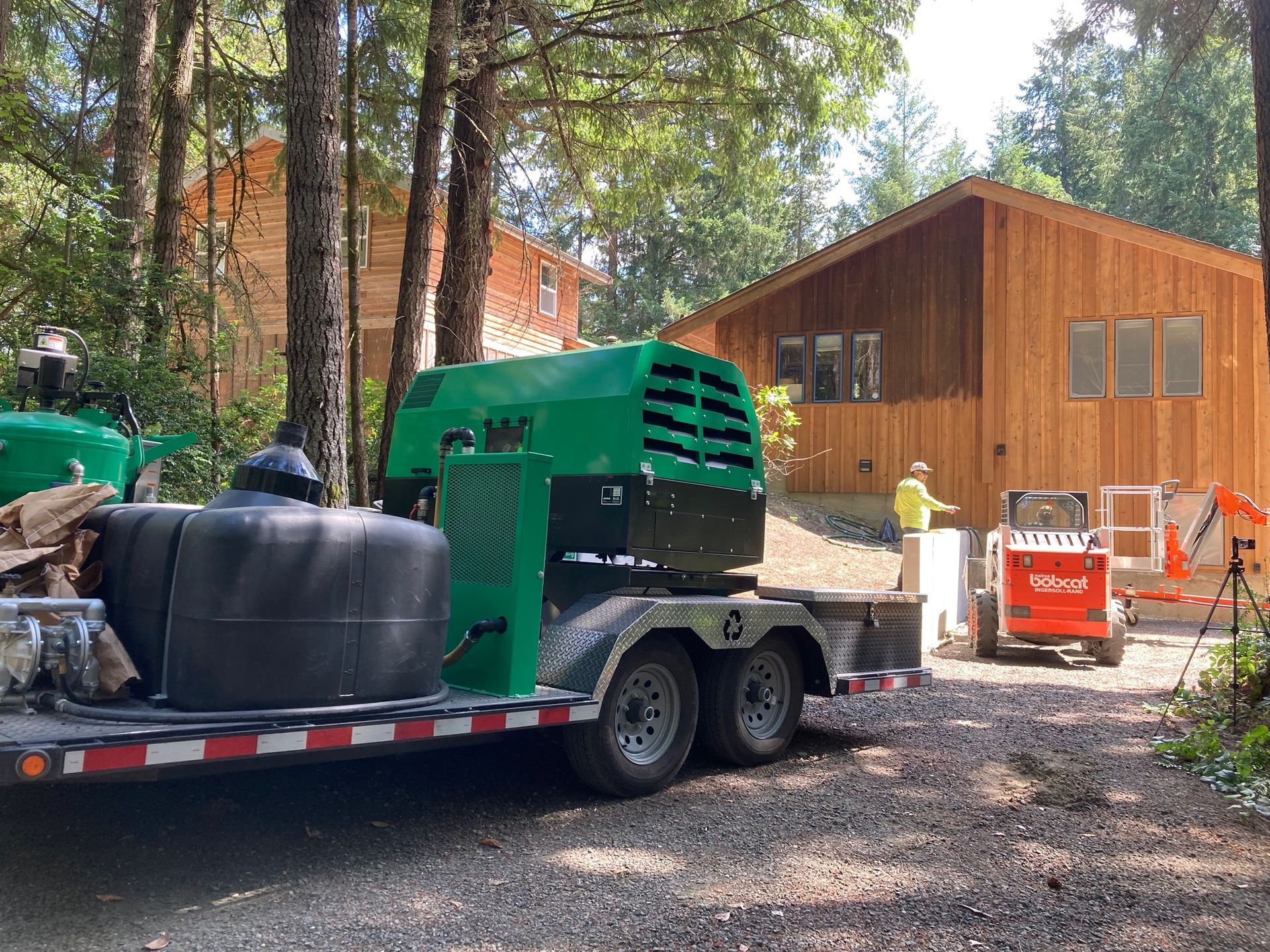 Construction site with equipment, two buildings in the background, a trailer with machinery, and a worker.