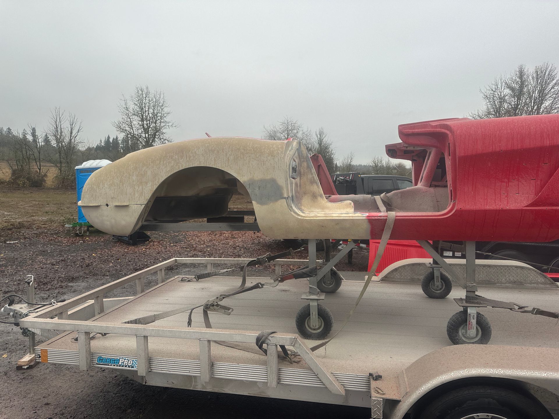 A red car body and fiberglass section on a trailer, outside, on a cloudy day.