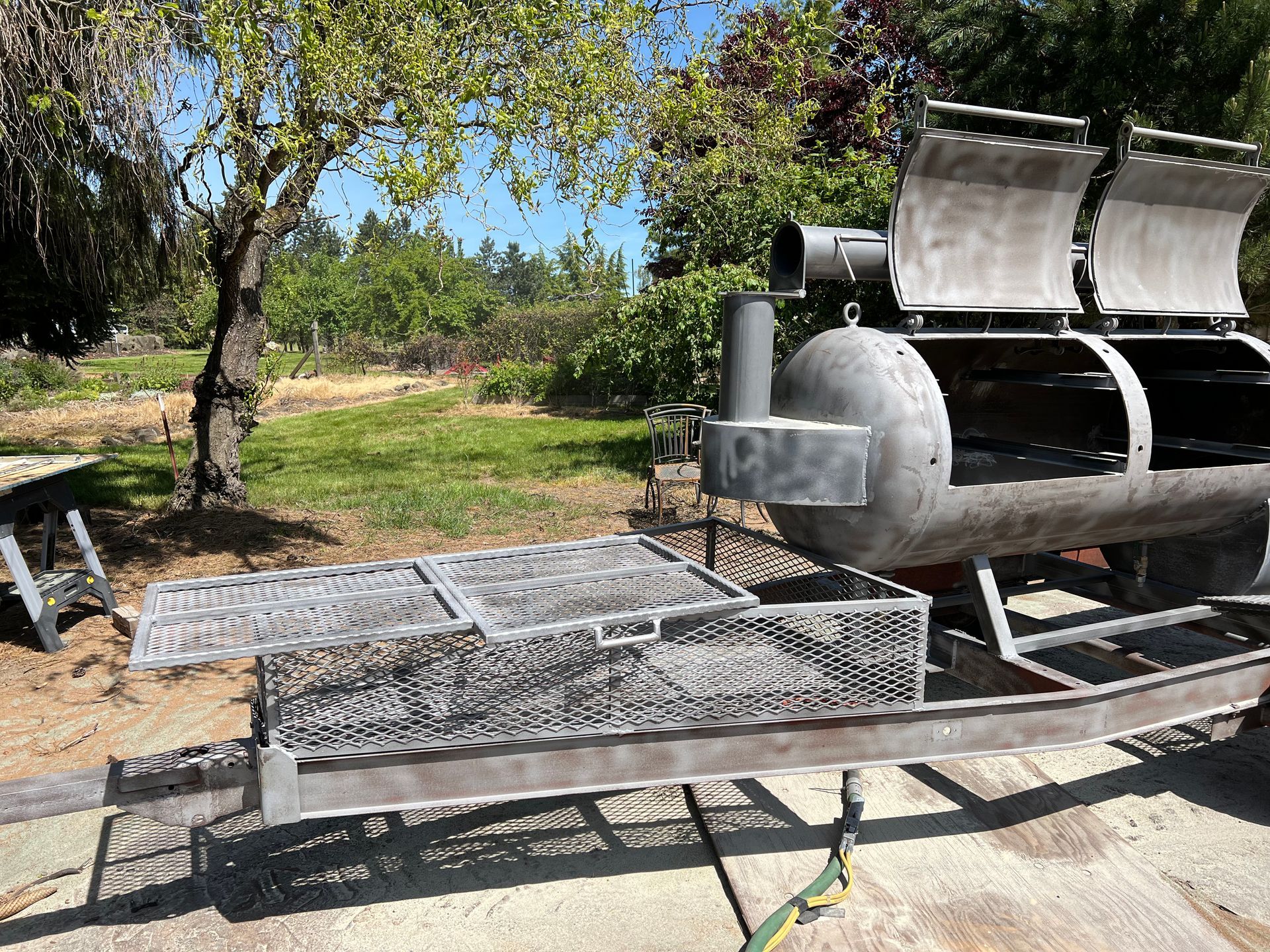 A large, metal barbecue smoker with two cooking chambers and a grilling surface, set on a trailer, outdoors.