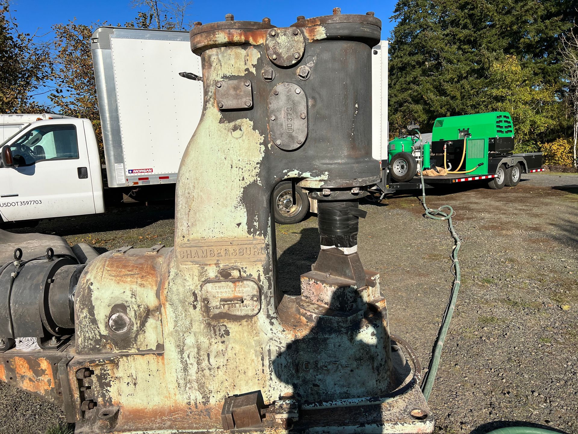 Large, old, industrial hammer outdoors; faded green and black with a truck, trailer, and trees in the background.