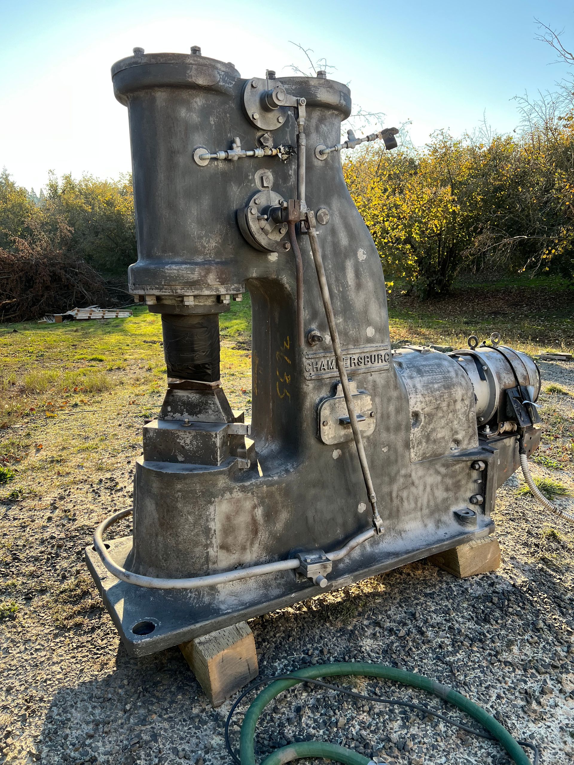 Large, gray industrial power hammer on wooden blocks in a grassy outdoor setting.