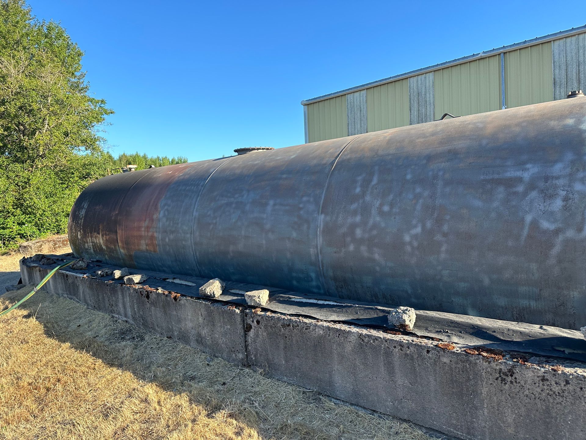 Large, rusted cylindrical tank on a concrete base, set outdoors near a building under a blue sky.