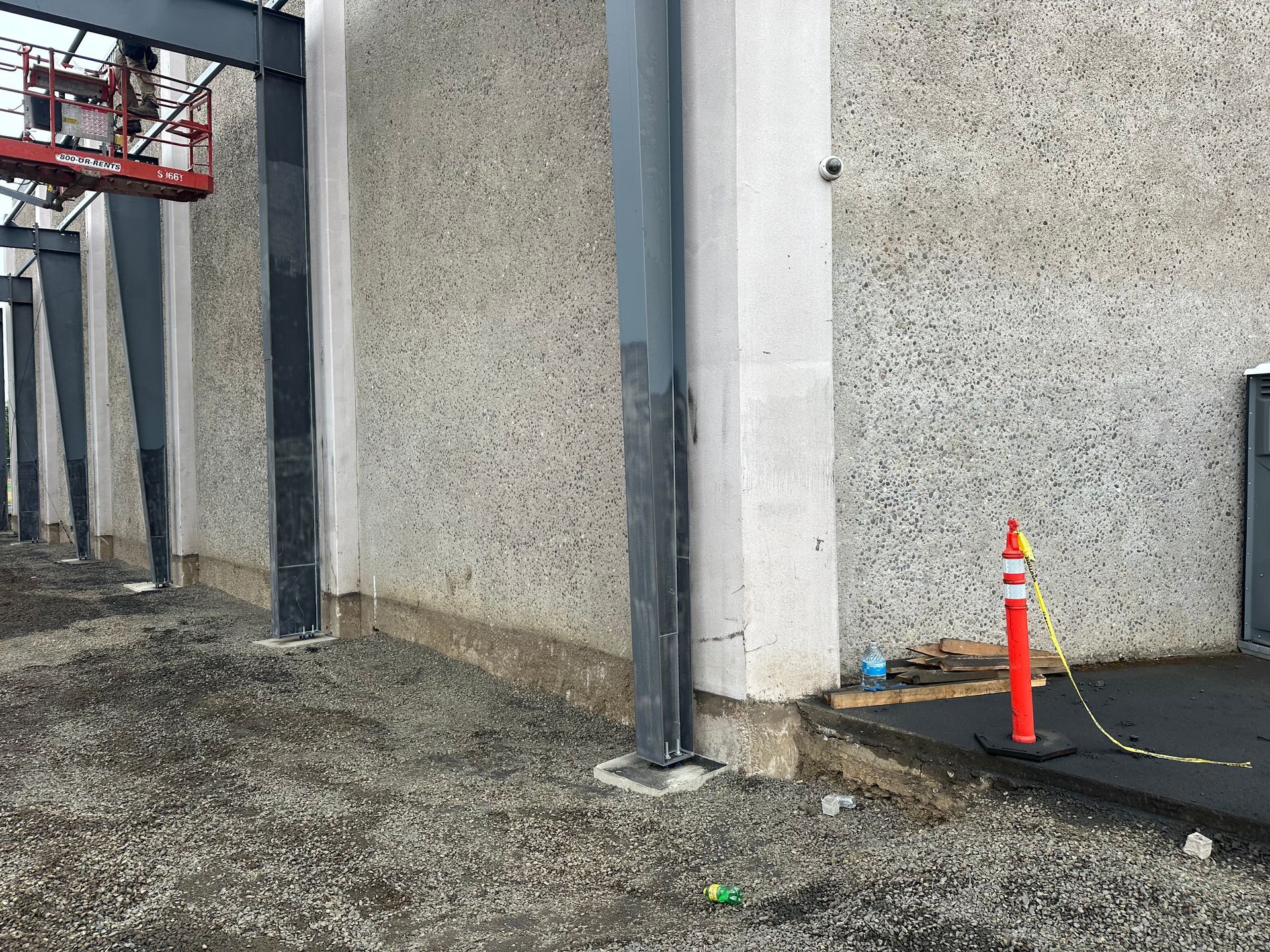 Exterior of a building under construction, showing concrete wall, metal beams, gravel, and an orange traffic cone.