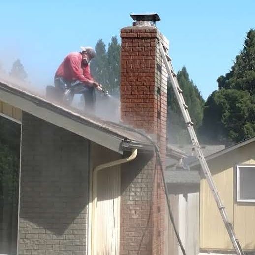 Man pressure-washing a brick chimney from a rooftop. A ladder leans against the chimney.