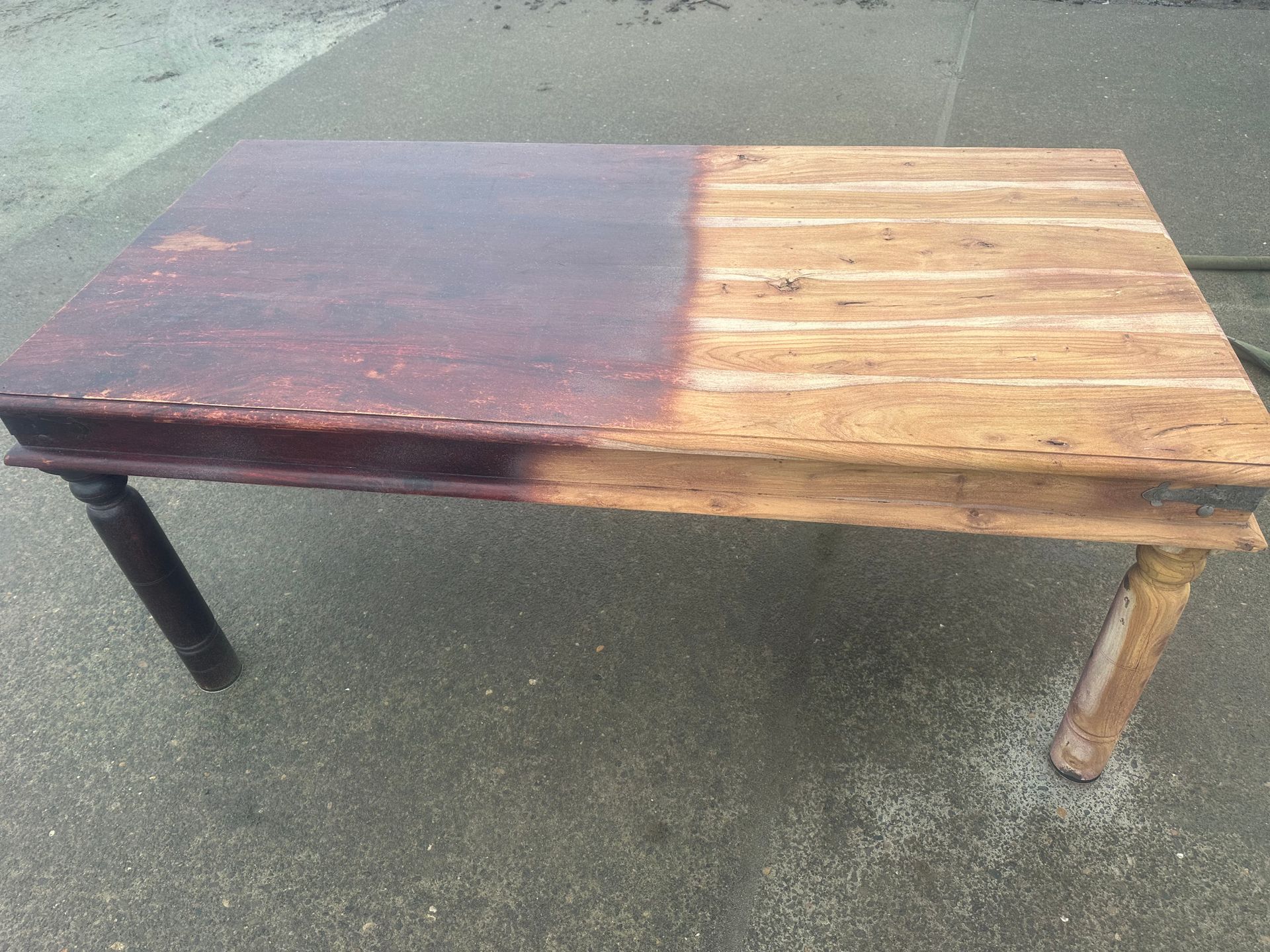 A wooden table, half stained dark brown and half unfinished light wood, resting outside.