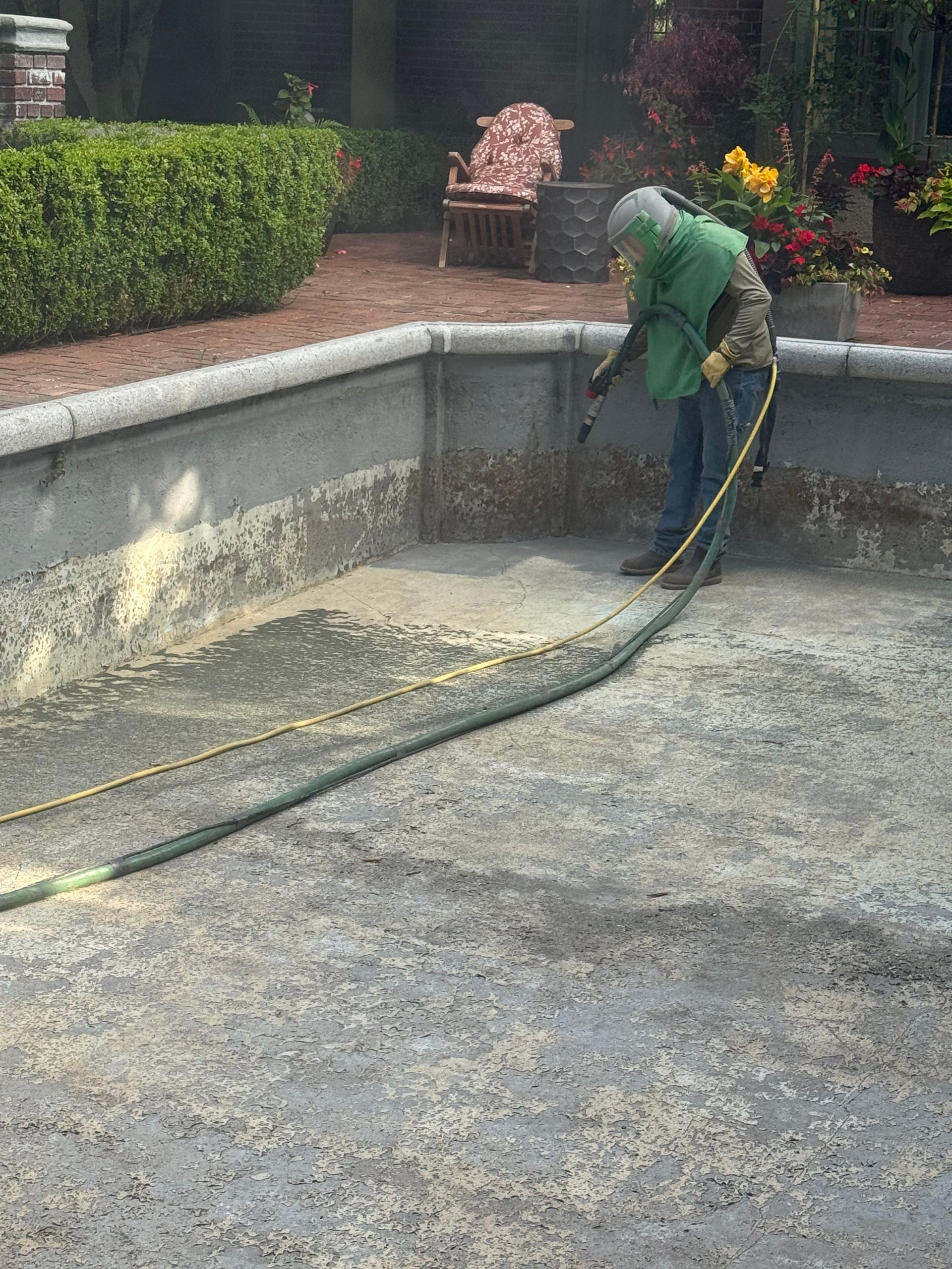 Person in protective gear sandblasting the inside of a concrete swimming pool.