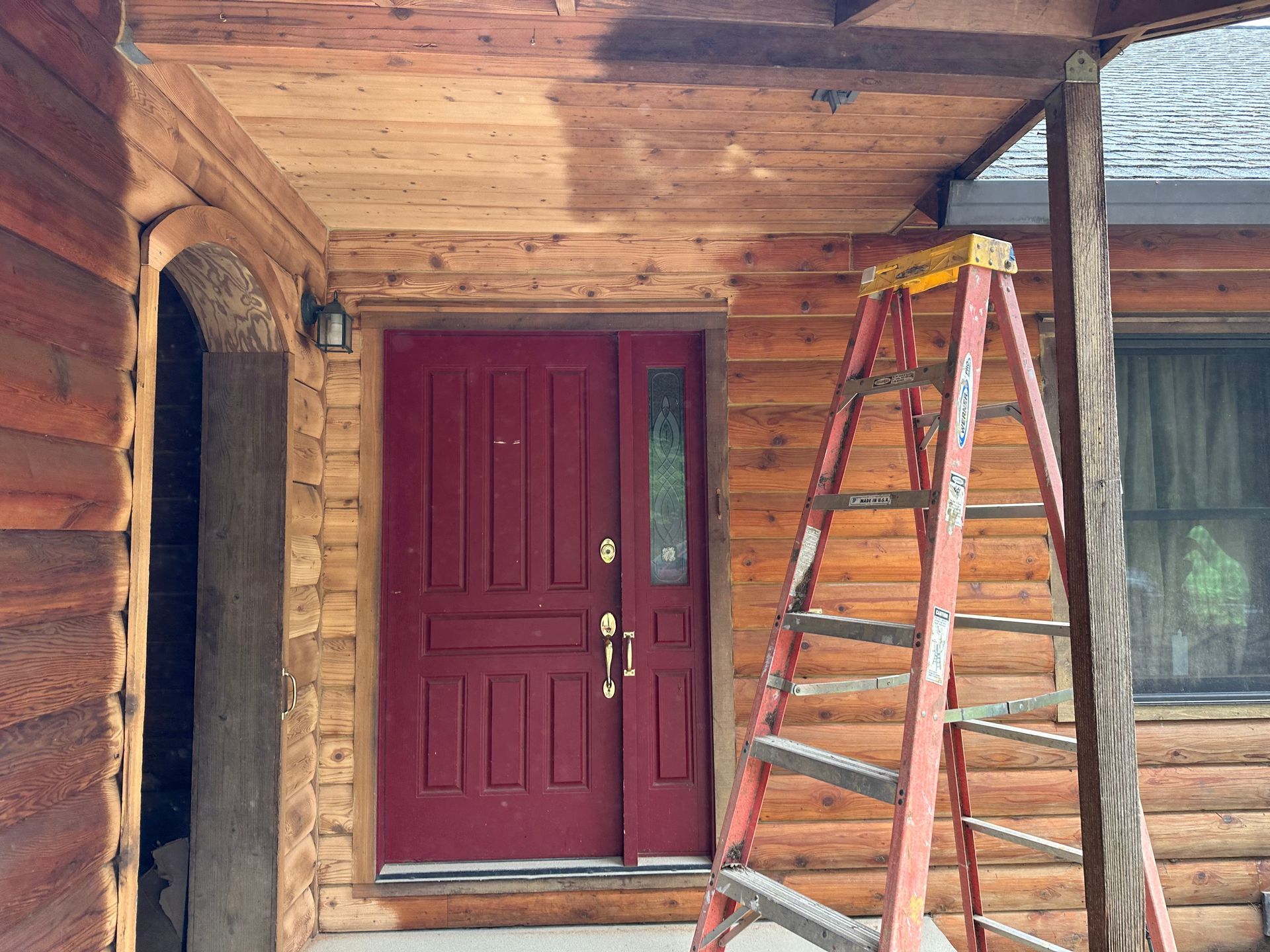 Red door on a log cabin porch; a ladder leans against the building.