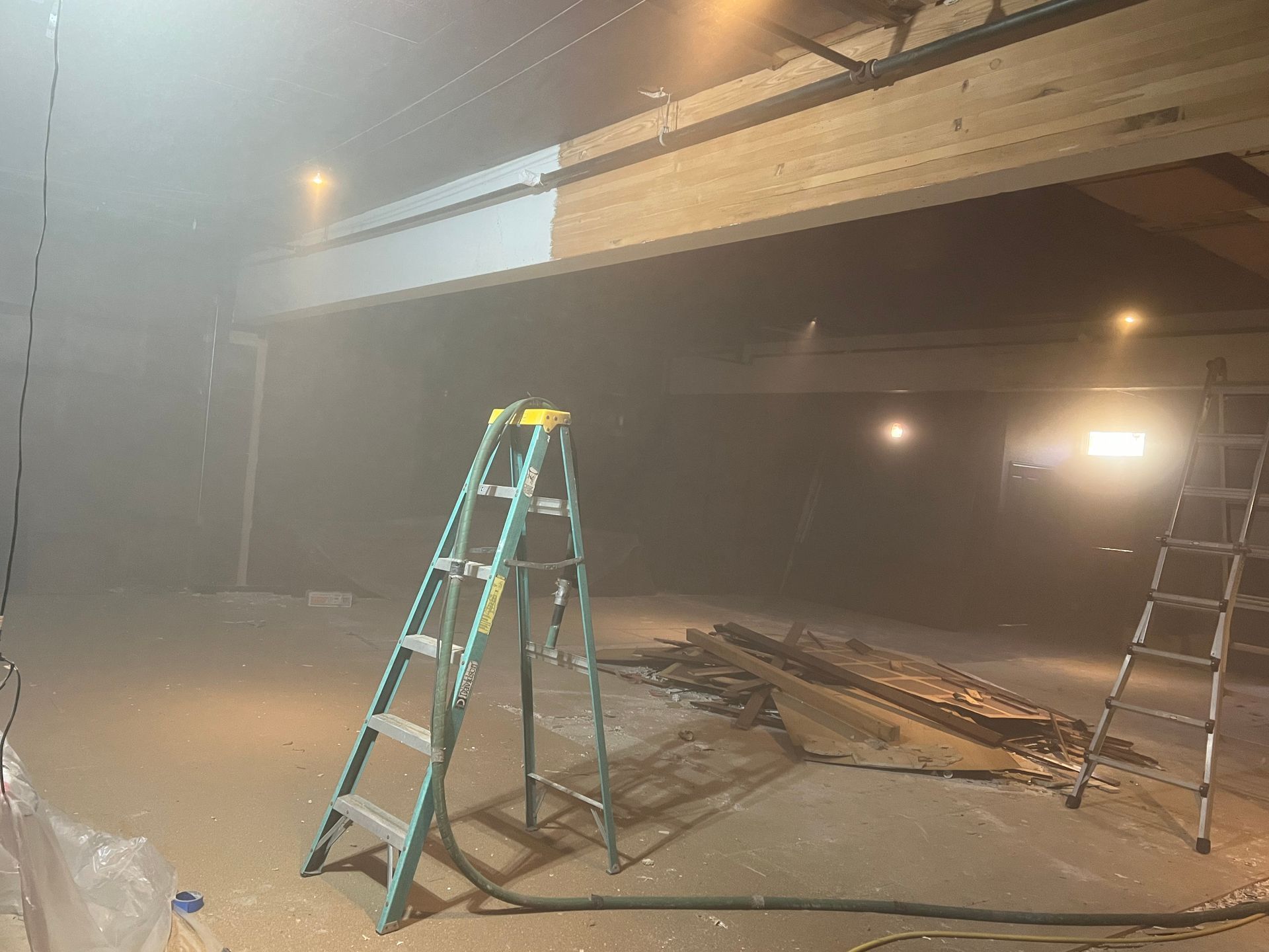 Construction site, dusty interior with ladder beneath a wooden beam being painted white. Another ladder is on the right.