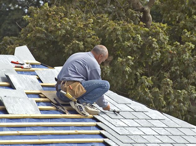 Worker crouches on a shingled roof, installing gray tiles near trees.