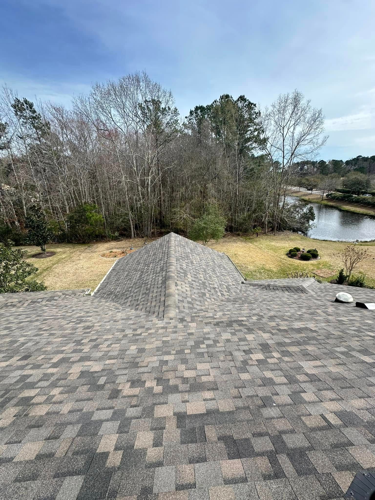 A high-angle view of a gray shingled roof with a pointed dormer, overlooking a grassy area and a body of water.