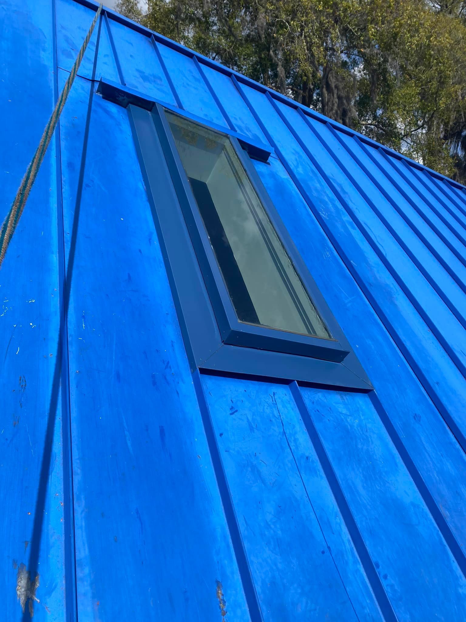 A rectangular skylight set into a bright blue metal standing-seam roof under a sunny sky with tree branches above.