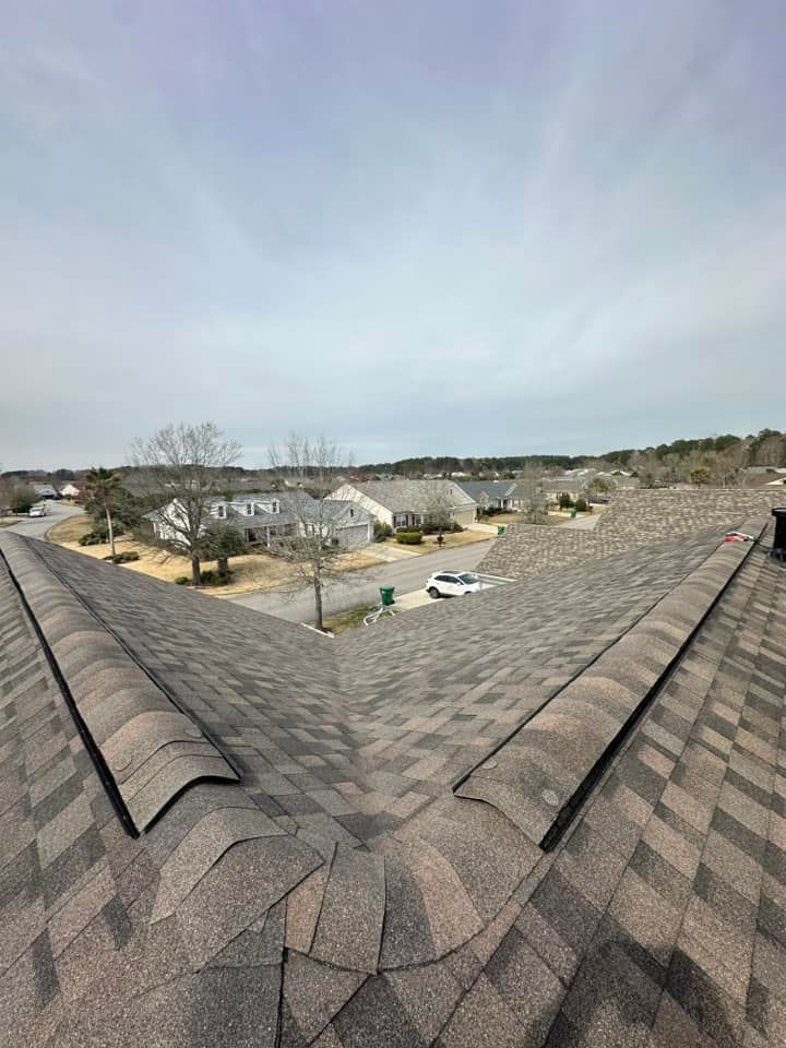 A high-angle view of a residential shingled roof looking down toward a suburban street with houses and trees.