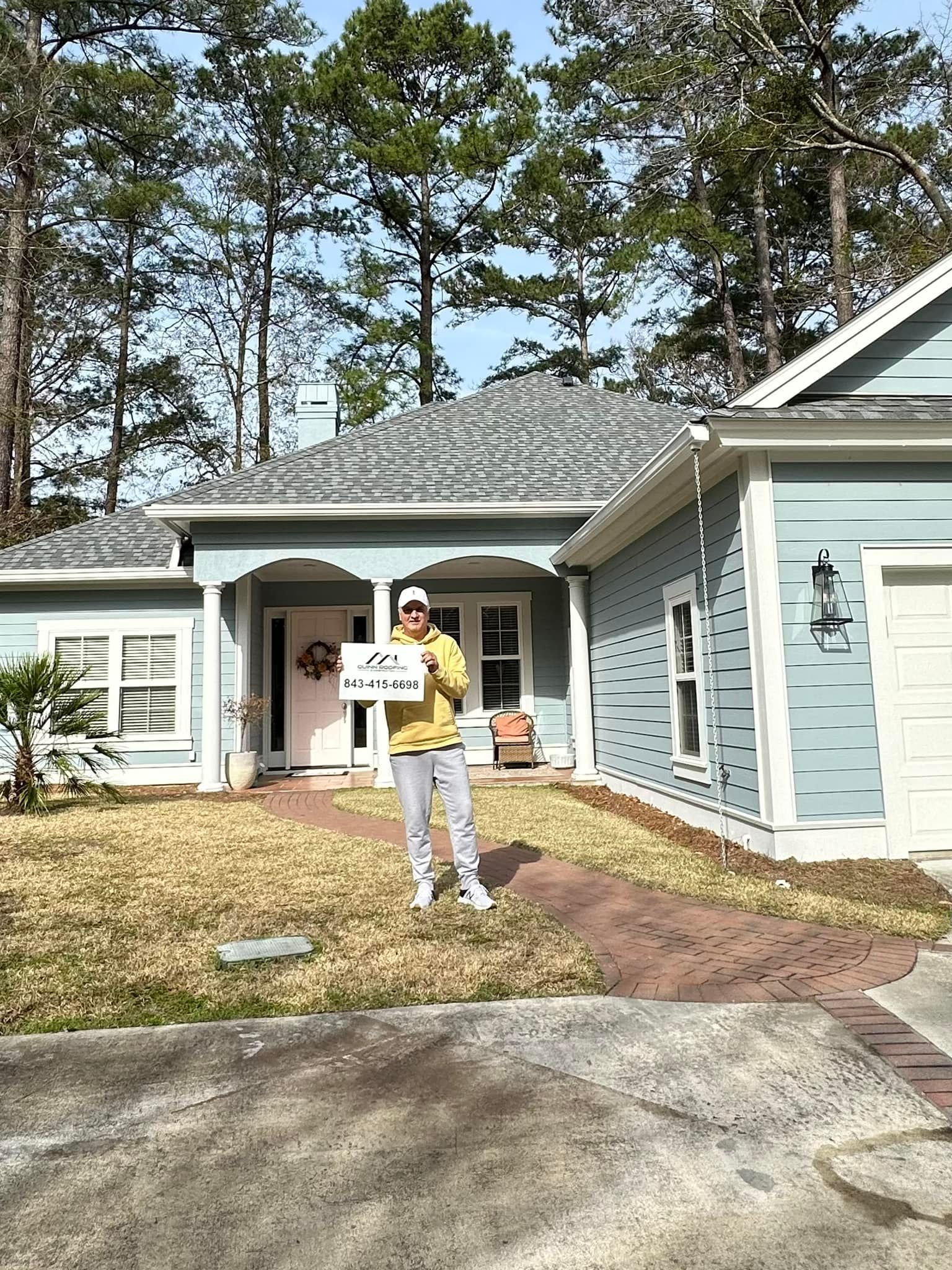 A person in a yellow hoodie stands in front of a blue suburban house holding a sign with text.