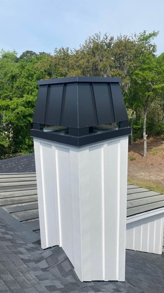 A white, board-and-batten chimney stack on a roof with a black, flared metal chimney cap against a treed background.