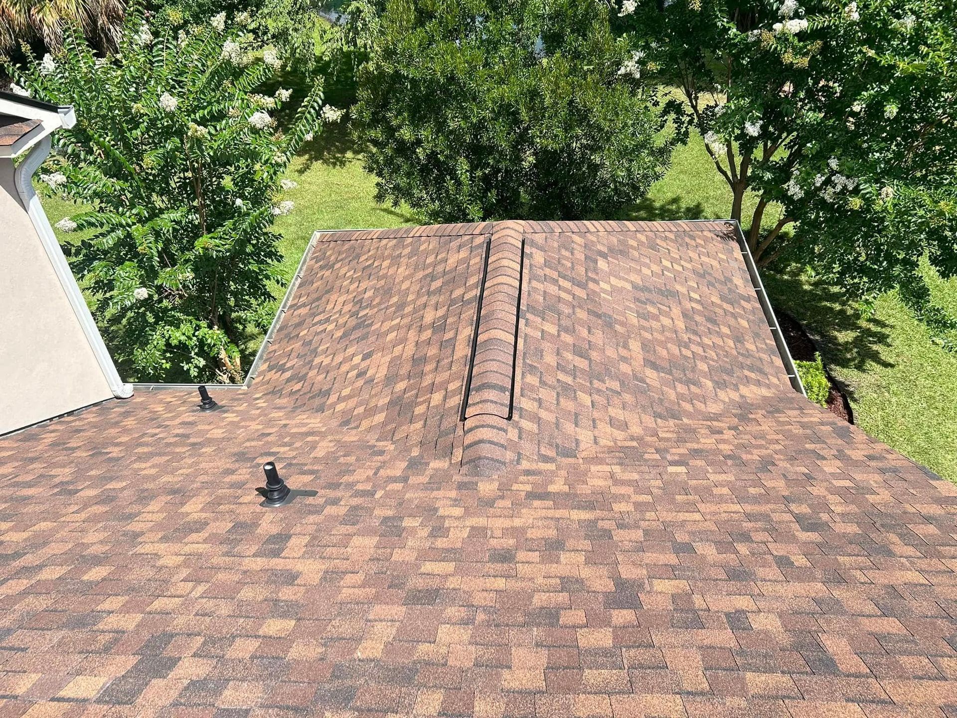 An overhead view of a brown shingled roof with a central ridge vent, surrounded by trees on a sunny day.