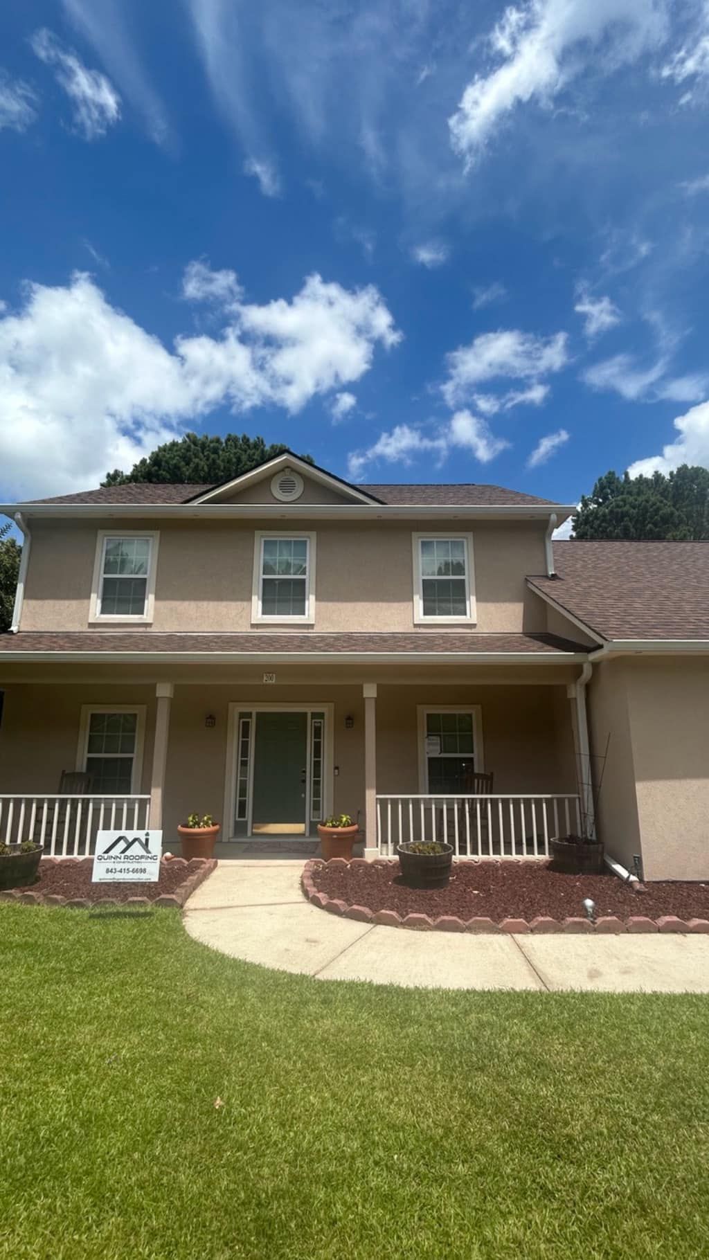 A two-story tan house with a dark roof and front porch, set behind a manicured green lawn under a bright blue sky.