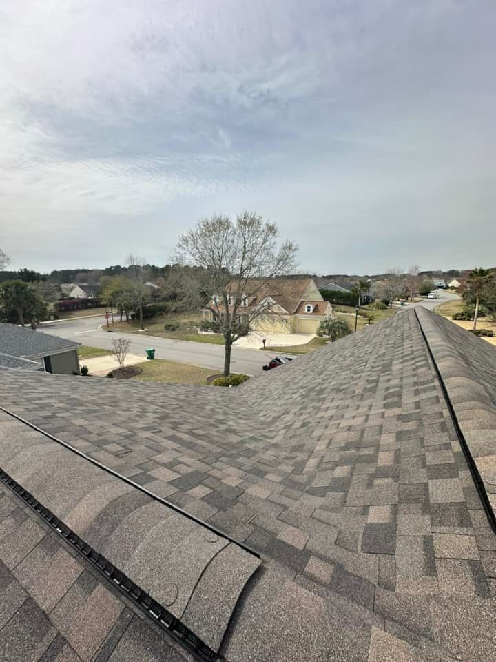 A high-angle view of a grey shingled roof looking out over a quiet suburban street with trees and houses.