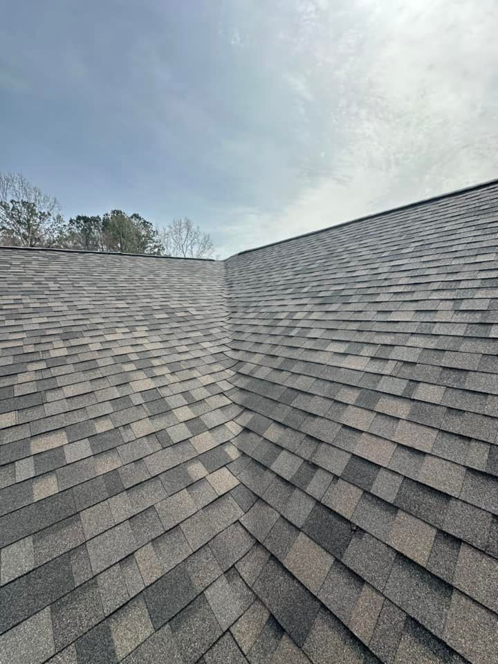 A valley on a gray, architectural shingle roof under a cloudy sky.