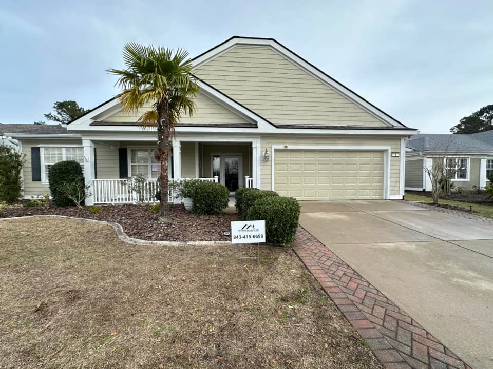 A single-story tan house with a two-car garage, white trim, a front porch, and a palm tree in the landscaped front yard.