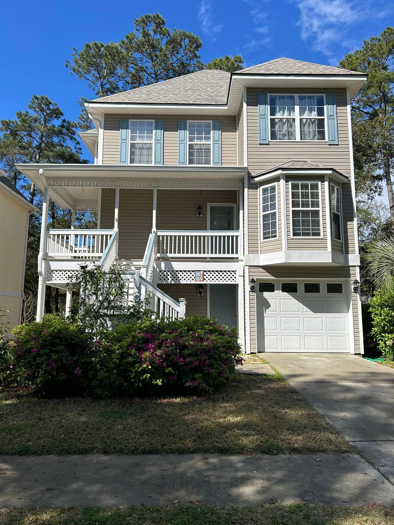 A two-story tan house with a front porch, white railings, a bay window on the second floor, and a one-car garage.