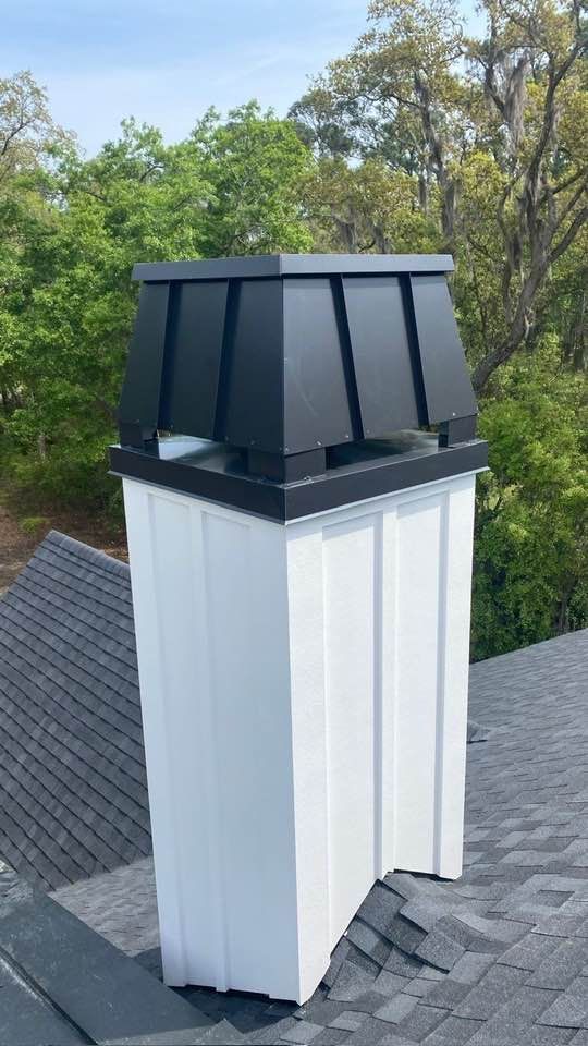 A chimney with a white board-and-batten base and a dark, flared metal cap sitting on a shingled roof against green trees.