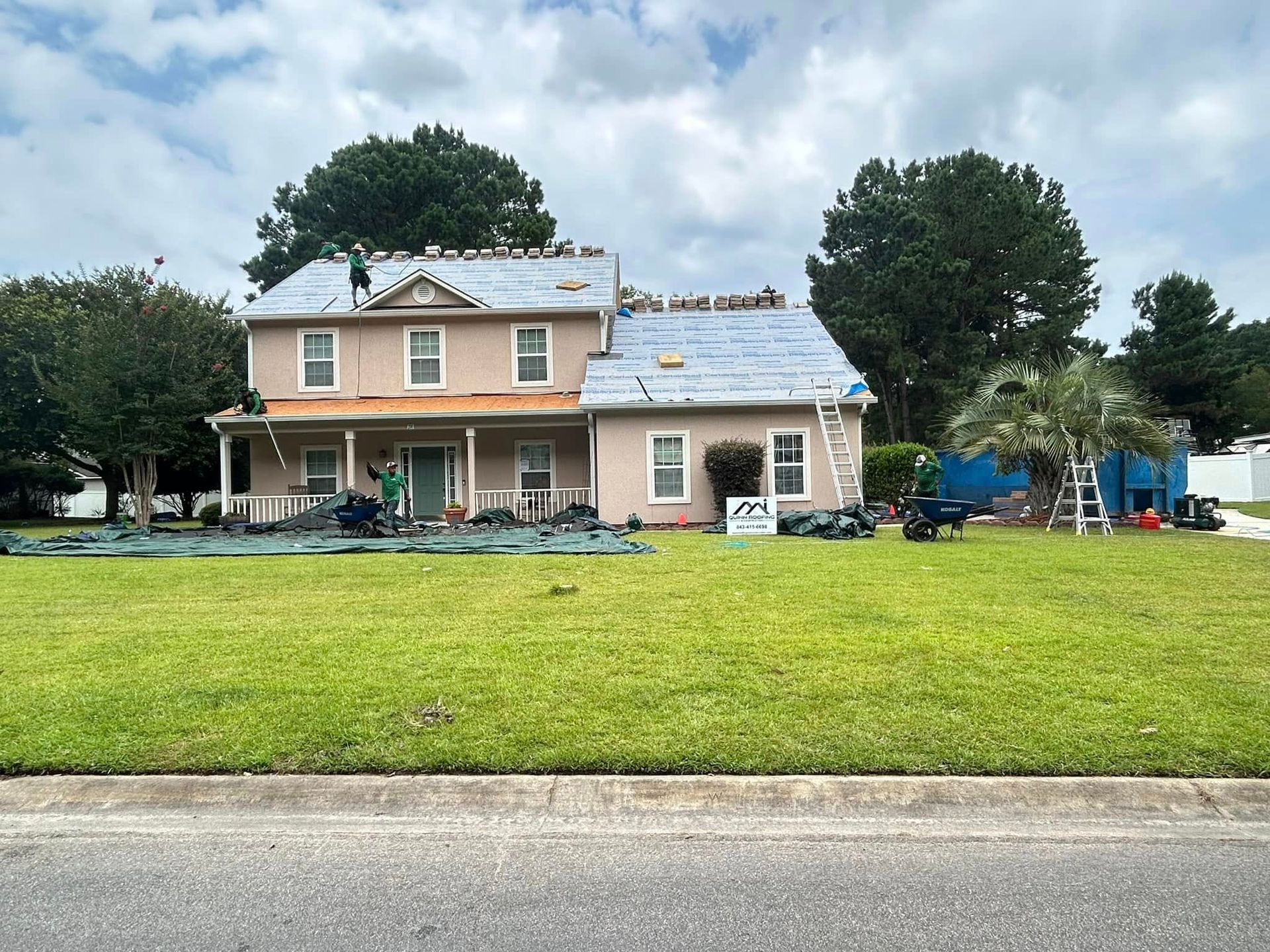 Workers replace the shingle roof on a two-story tan house with a large green lawn in front.