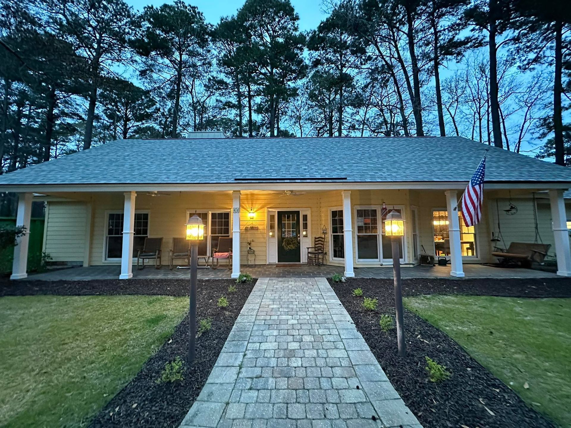 A light yellow house with a stone walkway and a wide front porch, set against a backdrop of tall pine trees at dusk.