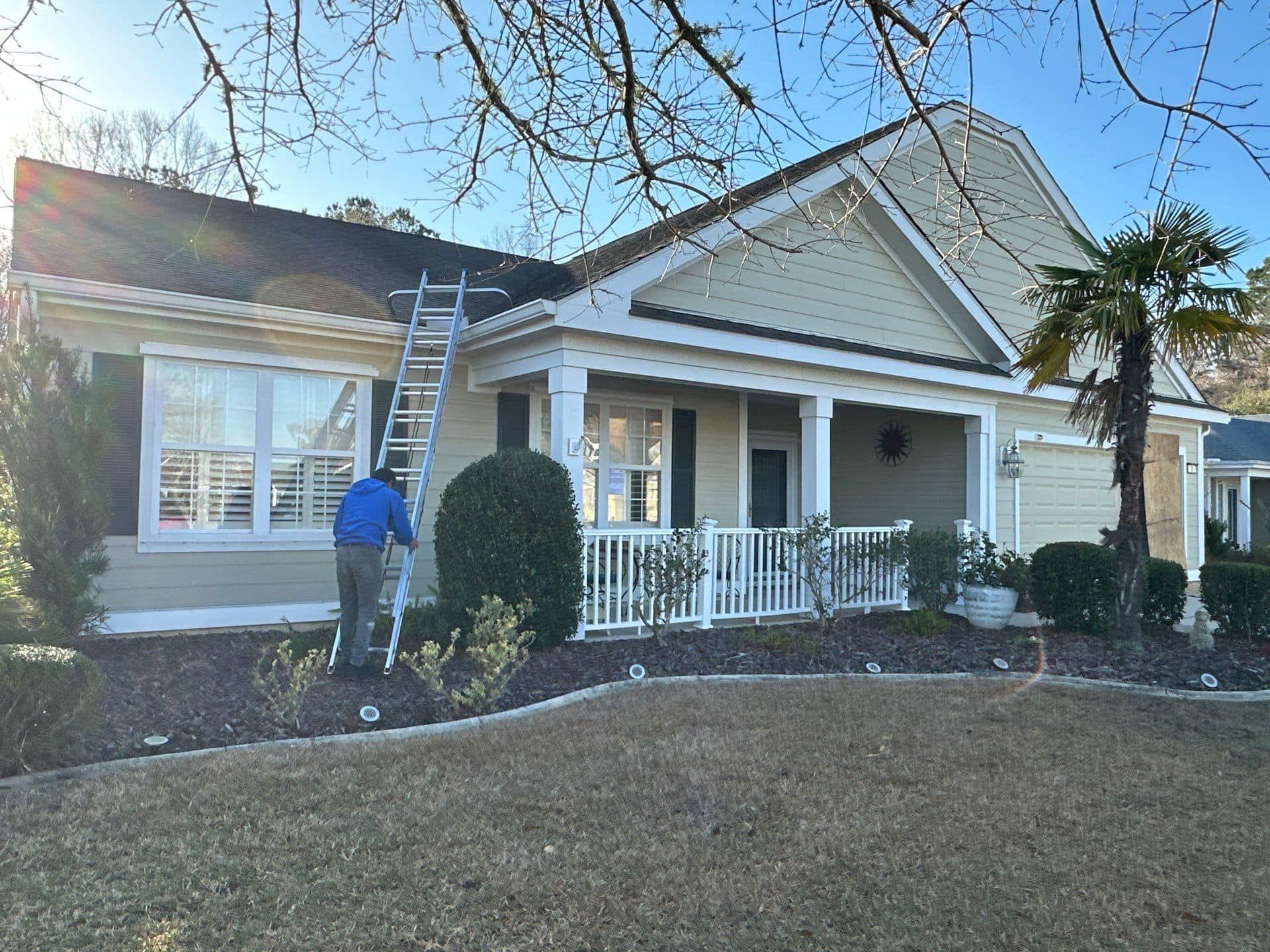 A person in a blue shirt climbs a ladder resting against the roof of a light yellow, single-story house.