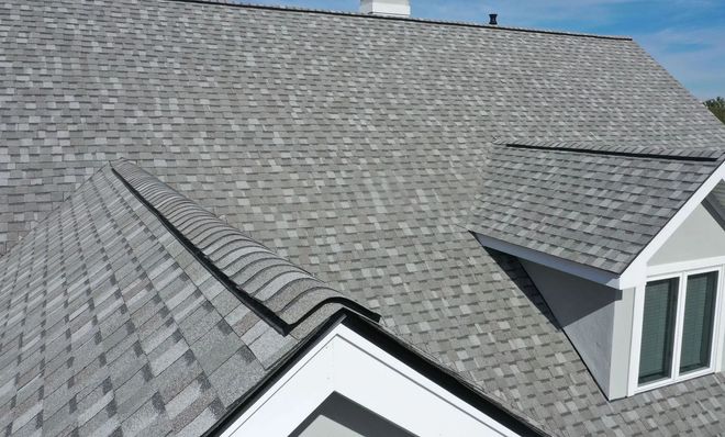 An aerial view of a gray asphalt shingle roof with a prominent dormer window and white trim against a blue sky.