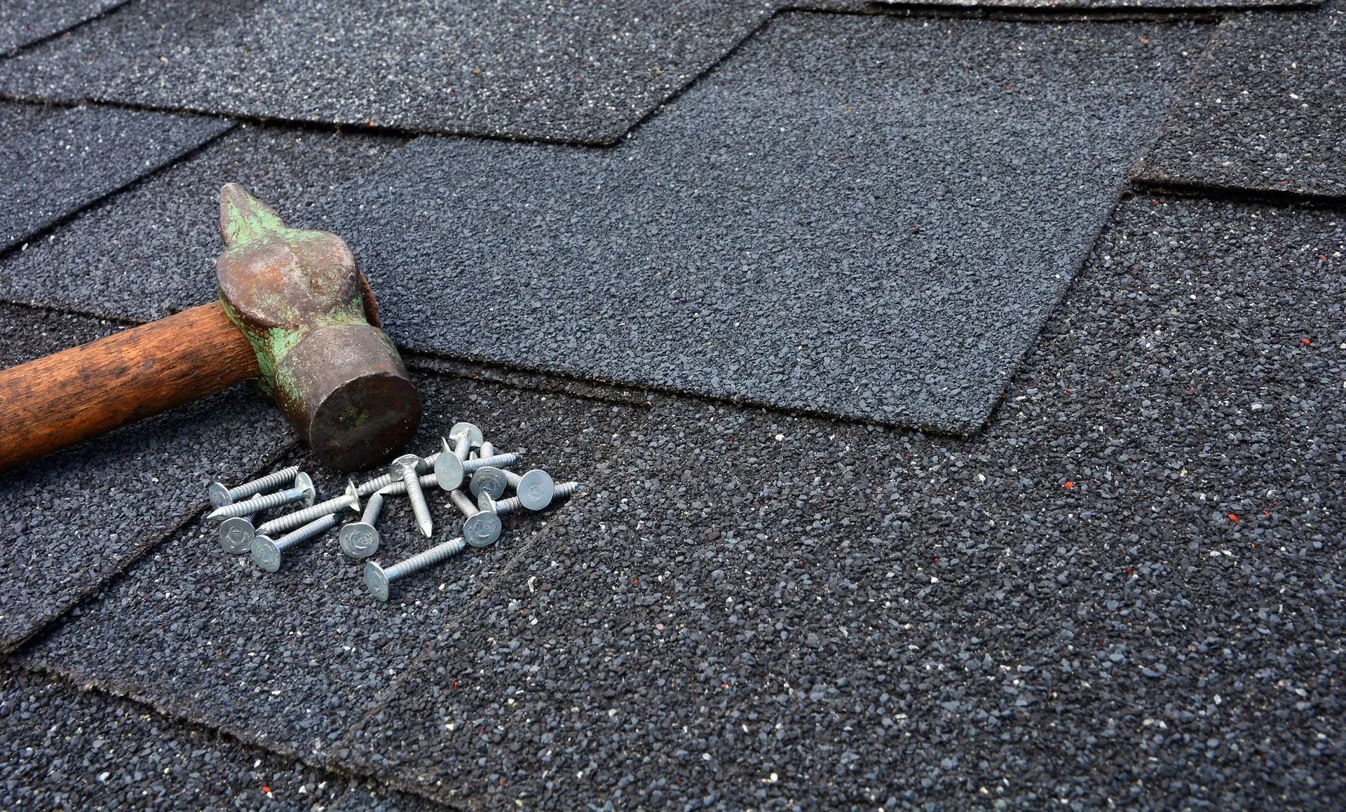 A weathered hammer lies on dark asphalt roof shingles next to a small pile of galvanized roofing nails.
