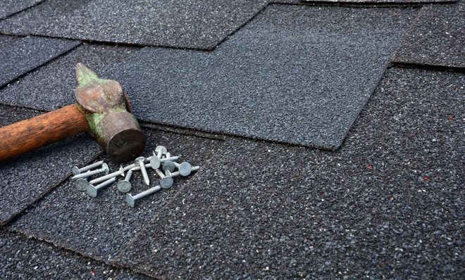 A weathered hammer lies on dark asphalt roof shingles next to a small pile of galvanized roofing nails.